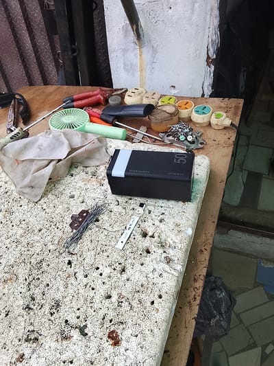 The image shows a collection of tools and electrical components scattered on a wooden surface and a textured white material. The objects include a pair of red-handled pliers, screwdrivers with green and black handles, a small green fan, a piece of cloth, a black power bank with "ACE ELEC" and "50" printed on it, and several electrical plugs and sockets of various colors, including yellow, green, and orange. There are also loose screws and wires, some of which appear to be part of a disassembled electronic device. The background is out of focus, showing a corrugated metal wall and a light-colored, textured wall. The overall impression is of a work area or a storage space for electronic repair or tinkering. The lighting suggests it is daytime, but the specific time and weather conditions are not discernible. The location is identified as Rumuepirikom, Nigeria.