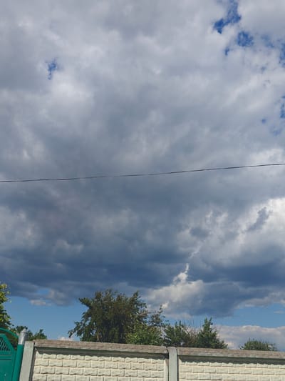 The image depicts a cloudy sky with patches of blue visible. A thin wire stretches horizontally across the upper half of the frame. In the lower half, a concrete fence with a light-colored, brick-patterned surface runs across the width of the image. To the left of the fence, a portion of a green metal gate is visible, along with a concrete post topped with a cylindrical light fixture. Behind the fence and gate, trees with green foliage are present. The scene appears to be an outdoor location, potentially a backyard or residential area in Kotelva, Ukraine, given the provided context.