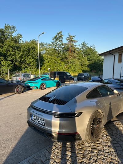 In Kerenite, Bulgaria, under a clear blue sky, a collection of Porsche vehicles is parked in an outdoor lot. The vehicles, all modern sports cars, are arranged in a row, with a prominent grey Porsche 911 Carrera GTS in the foreground, facing right. To its left, a bright teal Porsche 911 is visible, followed by a black Porsche on the far left. Behind these, a dark colored van and a dark grey SUV are parked. The scene is characterized by the presence of lush green trees and a building with a white facade and a dark roof on the right side of the frame. A street lamp stands tall in the background. The ground is paved with asphalt and cobblestones, with distinct shadows cast by the vehicles and trees. The license plate of the grey Porsche reads "CB 8887 XE".
