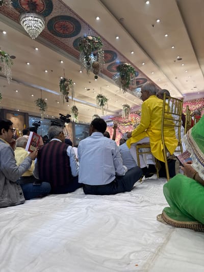This image depicts an indoor gathering at a large venue in Bidhannagar, India, possibly a banquet hall or community center, adorned for an event. The ceiling features multiple ornate crystal chandeliers and numerous hanging decorative elements composed of artificial white flowers and greenery. The ceiling itself is a mix of white panels and sections with patterned fabric in shades of pink, blue, and gold, featuring circular motifs and borders. Recessed lighting is visible throughout the ceiling.

Attendees are primarily seated on a white sheet or mat covering the floor. In the foreground on the left, a man wearing a grey jacket over a yellow shirt and blue jeans holds a small red-covered book. Next to him, from the back, a man in a maroon and black striped waistcoat over a light shirt and dark trousers is seated. Another man to his right, viewed from the back, wears a light blue checkered shirt and dark trousers, also seated cross-legged. On the far right foreground, a person clad in a vibrant green garment, possibly a saree, holds a small book.

In the mid-ground, a prominent man with grey hair is seated on an ornate golden chair, wearing a bright yellow traditional Indian kurta over white trousers. He is looking towards the left. Behind the seated attendees, professional video cameras on tripods are visible, indicating the event is being recorded or broadcast. The background shows bright, possibly decorative, lighting and elaborate backdrops with pink and gold patterns, suggesting a central stage or area of focus for the event. The individuals are generally attentive, with some holding books, implying participation in a shared activity like reading or listening.