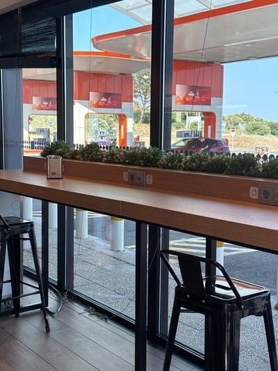 The image depicts an interior view looking out through a large window at a gas station, likely located in Oeiras, Portugal, given the prominent "Galp" branding on the gas station structures, which is a major Portuguese energy company. The window forms a long counter, made of a light-colored wood-like material, with two black metal stools placed in front of it. Mounted on the counter are electrical outlets and USB ports. A glass napkin dispenser sits on the counter near the left edge.
Outside the window, the gas station features several orange and white structures, with large banners displaying the "Galp" logo and advertisements. Gas pumps are visible, and a red car is parked at one of them, with a person leaning into the car from the passenger side. The environment appears to be sunny, with a clear blue sky. Lush green bushes are planted along the exterior of the window, forming a decorative border. In the foreground, on the left, a portion of another black metal stool is visible, and on the floor, electrical cords are tangled. The flooring inside the establishment appears to be dark wood planks.
