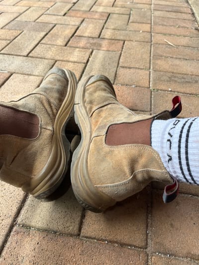 A pair of tan work boots, likely made of suede or a similar material, sit atop a brick surface. One boot is slightly overlapping the other. A white sock with black stripes and the word "RVCA" visible peeks out from the top of the boot. The boots appear worn with some dirt and scuffs, suggesting they've been used for work. The brick background is textured with various tones of brown.