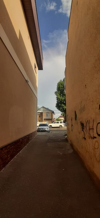 The image depicts a narrow alleyway between two buildings in Yessentuki, Russia. The building on the left is light beige with a brown brick base and a dark brown roof overhang. The building on the right has an orange-beige facade with graffiti. A paved pathway runs through the alley. In the background, two cars are visible: a silver sedan in the foreground and a white SUV parked behind it. A two-story building with a light brown facade and white trim, along with a large green tree, are also present in the background. The sky above is blue with scattered white clouds.