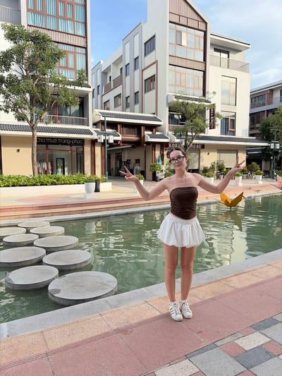 A young Caucasian woman with dark hair pulled back and wearing glasses stands on a paved walkway next to a body of water with circular stepping stones. She is wearing a brown strapless top and a white ruffled skirt, paired with white sneakers. Her arms are raised, and her hands are making peace signs. Behind her, there are modern, multi-story buildings with balconies and large windows. The environment appears to be a commercial or residential area with some greenery and decorative elements. The signage on the buildings suggests a commercial establishment, possibly a shop. The scene is outdoors, during daylight, with a clear sky. The location is identified as Nghĩa Trụ Commune, Vietnam.