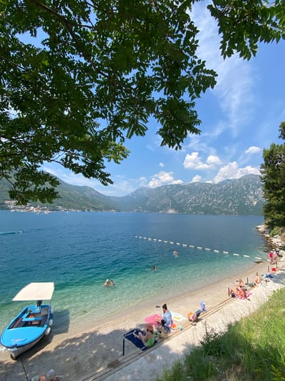 The image depicts a serene summer scene at a beach in Perast, Montenegro. A bright blue boat with a canopy is docked near the shore, its hull resting on the sandy beach. The crystal-clear turquoise water gently laps against the shore, revealing the sandy bottom. Several people are visible enjoying the water and the beach. Two individuals are swimming in the clear water, while others are sunbathing on lounge chairs or towels spread on the sand. A rope with buoys demarcates a swimming area in the sea. In the background, lush green mountains rise majestically, their slopes dotted with buildings, suggesting the picturesque town of Perast. The sky above is a vibrant blue, with scattered white clouds adding to the idyllic atmosphere. The scene is framed by the leafy branches of a large tree in the foreground, casting dappled shadows on the beach.