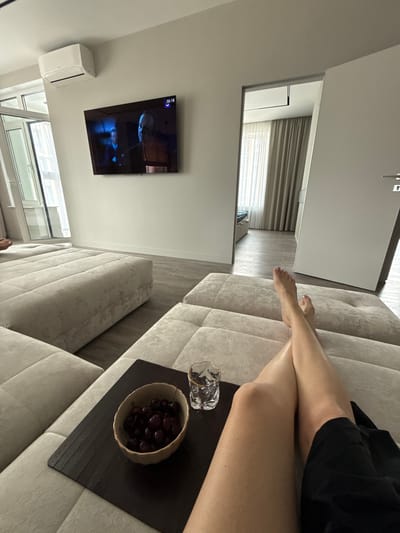 The image shows a person's legs and feet resting on a beige upholstered sofa in a modern apartment. The person is wearing a black garment. In front of them, on a dark placemat, is a small bowl of cherries and a glass of water. The legs are crossed at the knees, and the feet are bare. A television is mounted on the wall, displaying a program with a person's face on screen. An air conditioning unit is visible below the TV. The room has light-colored walls and wooden flooring. A large window with blinds is also visible. The overall impression is one of relaxation and leisure. The time on the TV screen reads 16:18. The location is Dolgoprudny, Russia.