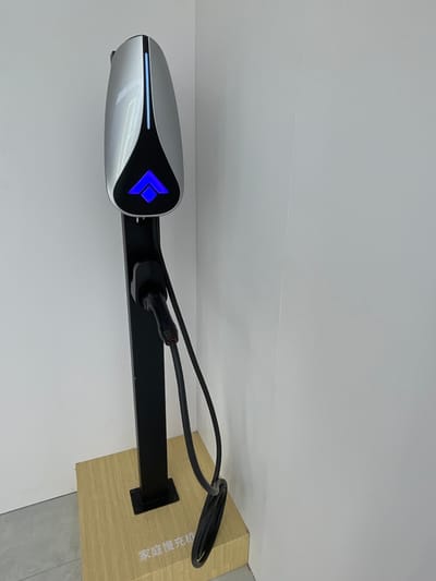 The image shows an electric vehicle charging station located in Hangzhou, China, at the coordinates 30.272 latitude, 120.416 longitude. The station is mounted on a black metal pole with a white charging port, featuring a blue illuminated arrow symbol, attached to the top.  The charging cable, coiled on the floor, is plugged into the station. The station is positioned on a wooden platform, featuring Chinese text in the lower right corner.  The station is mounted on a white wall in a room with grey tile flooring.
