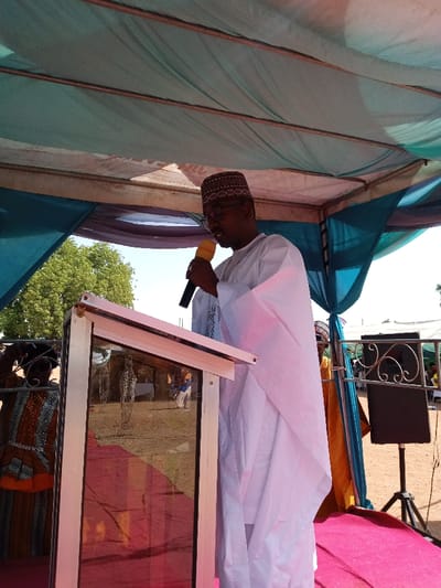 The image shows a man, wearing a white robe and a red and yellow cap, speaking into a microphone behind a podium. He is standing on a red and white carpet, under a blue and white canopy. The podium is made of metal and has a glass pane. There is a black speaker on a tripod in the background. There is a partial view of another person wearing a yellow and black robe to the left. The scene is likely outdoors and appears to be a public event. The location indicated by the coordinates (latitude: 10.116, longitude: 13.046) is Zum, Nigeria.