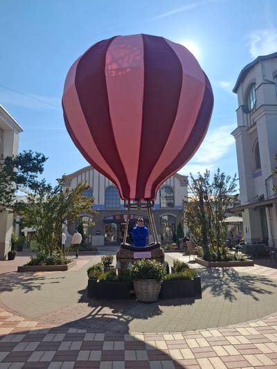 The scene depicts a vibrant outdoor pedestrian plaza at Ingolstadt Village, Ingolstadt, Germany, under clear blue skies with direct sunlight. A large, non-operational hot air balloon display, featuring alternating dark red and light pink vertical stripes, occupies the central foreground. Its dark brown wicker basket is encircled by dark planter boxes containing green foliage. A male subject, wearing a blue long-sleeved top and sunglasses, is positioned inside the balloon's basket with his arms crossed.
The plaza's ground is paved with interlocking bricks in geometric patterns of tan, beige, and reddish-brown. Distinct shadows from trees and the hot air balloon are cast across the pavement, extending from the left and center. Surrounding the plaza are several multi-story buildings with light-colored facades, large arched windows, and multi-paned glass. These structures house retail establishments, identifiable by signage such as "BOSS" and "COFFEE."
Multiple small, leafy trees are planted in dark rectangular planters throughout the plaza. In the mid-ground left, two male subjects are observed walking away from the camera. To the right and deeper in the background, several individuals are seated at outdoor tables under umbrellas or awnings, presumably at café or restaurant areas. The overall environment suggests an upscale retail and leisure destination.