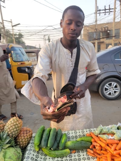 A vendor in a light-colored robe counts money from a black bag, near a display of fresh produce. Carrots, cucumbers, pineapples, and cabbages are neatly arranged on a patterned tablecloth. A car's wheel and a yellow tricycle are visible in the background, suggesting a street scene. Overhead power lines crisscross the sky.