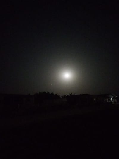 A nighttime scene with a bright moon dominating the sky. Below, a dark landscape reveals silhouettes of trees and a building with light emanating from its windows. The ground is obscured in darkness, with minimal details visible. A slight haze surrounds the moon, softening its light.