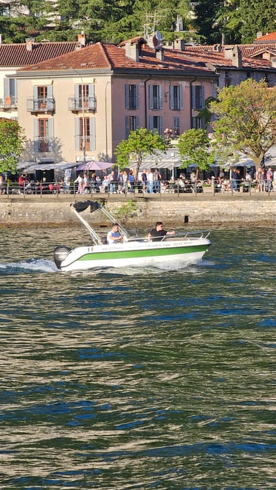 Here's a concise description of the image:
The image shows a waterfront scene in Como, Italy. A small motorboat, marked "RENT BOAT", is close to the shore. Two people are aboard the boat. A stone embankment runs along the water's edge, with numerous people sitting and standing along it, many appearing to be at outdoor restaurants or cafes. Buildings with tiled roofs and shuttered windows stand adjacent to the embankment. The water has ripples, and trees are visible among the buildings and along the waterfront. The overall scene suggests a sunny, tourist-oriented area.