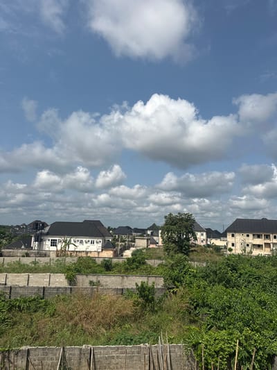 The image displays an outdoor daytime scene in Obinze, Nigeria, depicting a developing residential area under a blue sky with numerous scattered cumulus clouds. The foreground comprises an uncultivated area with multiple courses of uncompleted concrete block walls, interspersed with dry grasses, dense green foliage, and several vertical wooden stakes. Moving into the midground, additional concrete block walls form boundaries, behind which lush green vegetation and a prominent deciduous tree are visible. The background reveals a residential settlement consisting of numerous structures, primarily two-story buildings featuring dark, pitched roofs. A distinct white two-story building with multiple exterior air conditioning units and an elevated water tank is situated on the left side of the background. To its right, a multi-story building with an exposed, unfinished blockwork facade stands. Further back, more residential units with dark roofs are observable, connected by unpaved ground and interspersed with additional trees. No individuals or active engagements are depicted.
