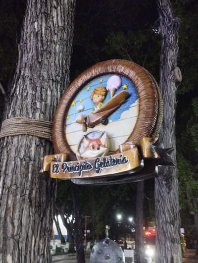 The image shows a sign for "El Principito Gelateria" (The Little Prince Ice Cream Shop), mounted on a tree in Valencia, Venezuela. The sign is round with a wooden frame, depicting The Little Prince in a plane, a fox, an ice cream cone, and stars. The sign is attached to the tree with rope. In the background, there are more trees and street lights, suggesting a park or urban green space. A spherical statue of the Little Prince can also be seen at the bottom. There are also cars in the background.