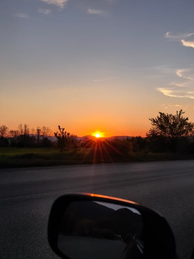 The image shows a sunset view from a vehicle, likely a car, with a side mirror visible in the foreground. The setting sun is centered in the background, positioned between trees and hills. The sky transitions from orange near the horizon to a lighter blue higher up with scattered clouds. A road surface is visible between the viewer and the landscape. The reflection in the side mirror shows a dark image of what seems to be the same landscape. The scene suggests the location is outside the city of Sapareva Banya, Bulgaria as it captures a rural or countryside environment with trees, hills, and a sunset.
