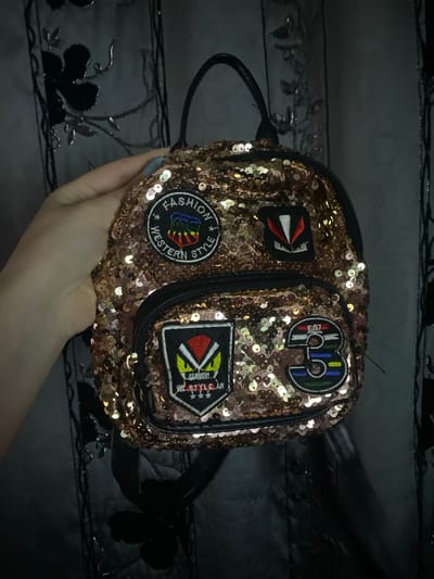 A person's hand holds a small, sequined bag in a dimly lit indoor setting. The bag is covered in shimmering rose gold-toned sequins and features several embroidered patches.

The visible patches include:
*   A circular patch with the text "FASHION STYLE WESTERN" surrounding a colorful, stylized emblem.
*   A shield-shaped patch displaying a red triangle, white arrows, and text that reads "WE STYLE GARMENT" and likely "HEARTWEAR."
*   Another patch, somewhat circular, features "EA7" and "SWIM" along with abstract shapes in green, blue, and red.
*   A smaller, angular patch with white and red design elements is also visible.

The hand, with light skin and light blue/grey nail polish, holds the bag against a dark, subtly patterned fabric background, possibly upholstery or a blanket. The background pattern appears to be floral or abstract, with some reflective elements. The overall lighting is dim, enhancing the sparkle of the sequins on the bag. The activity depicted is simply the presentation or display of the bag. The scene is described as being in Verkhnevesyoloye, Russia.