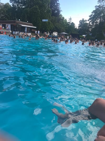 The image shows a crowded outdoor swimming pool, likely the "Anneliese Pohl Allee" public pool in Hattersheim, Germany, on a sunny day. The water is a vibrant turquoise blue and appears to be in motion with ripples and splashes. A large group of people of various ages are in the water, swimming, standing, and interacting. Some are closer to the foreground, while others are further back, towards the pool's edge.

On the left side of the pool, a white building with a brown roof is visible, appearing to be a changing room or concession stand. It has windows and a striped awning. Several people are standing on the deck near this building, and some are in the water close to the edge. In the background, lush green trees surround the pool area, suggesting a park-like setting. The sky above is clear and blue.

In the lower right corner of the image, the legs and feet of a person are visible, partially submerged in the water, indicating the viewpoint of the photographer. The overall impression is one of a lively and popular recreational area.