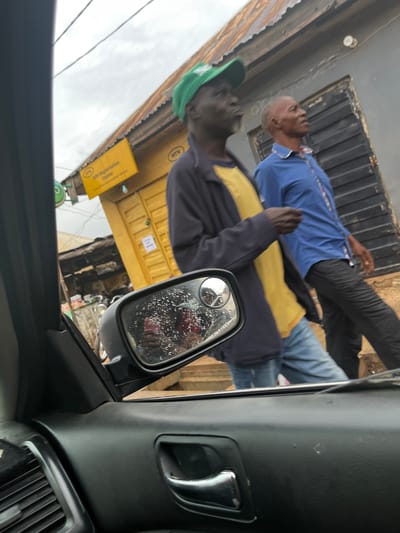 Two men are visible in the image, seen from the perspective of someone inside a vehicle. One man in the foreground wears a dark jacket over a yellow shirt and a green baseball cap. He is looking towards the left side of the frame. Behind him, another man is wearing a blue shirt and dark pants. He is looking upwards. Both men appear to be outdoors.

In the background, there is a building with a yellow facade. A sign with text is visible on the yellow building. One sign says "MTN". Another yellow sign has text that includes "SIM Registration Center". There are also some metal shutters on the building. The side mirror of the vehicle is visible in the foreground, and it has water droplets on its surface, suggesting it might be raining or has recently rained. The sky appears overcast, indicating a cloudy day. The scene appears to be a street in Sabojibi, Nigeria, given the contextual information.