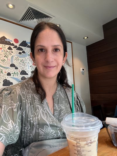 The image shows a woman with dark hair and brown eyes sitting at a table in what appears to be a Starbucks in San Isidro, Peru. She is wearing a green, floral-patterned blouse and pearl earrings. In front of her is a plastic cup with a clear lid and a green straw, containing a light-colored beverage. The cup has handwritten text on it, including the name "Ariana". In the background, there is a wall with a mural depicting mountains, trees, and a red sun, as well as a wooden panel. A white air vent is visible on the ceiling.