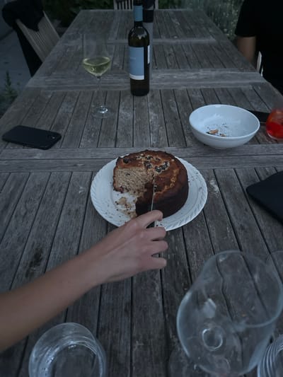 A wooden table, likely outdoors, displays a half-cut cake on a white plate, a hand holding a knife cutting it, along with a wine bottle, wine glass, bowl with cigarette butts, a drink with an orange slice, a cell phone, and empty glasses. The location is presumed to be Cambridge, United Kingdom. No people are fully visible. The general environment appears to be casual dining.