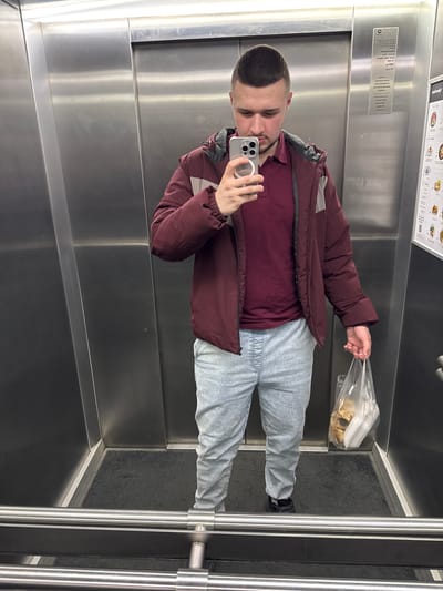 A man is taking a selfie in an elevator. He is wearing a maroon jacket over a maroon shirt and light gray sweatpants. He is holding a plastic bag containing an object, possibly food. The elevator is metal, with a black floor and metal handrails. There is a sign on the wall in the background. The location is inferred to be Bydgoszcz, Poland, based on the prompt's instruction.