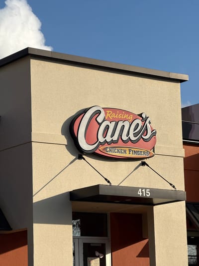 A Raising Cane's restaurant sign is affixed to the building's exterior, with "415" displayed on the awning above a window. The restaurant's facade features a light beige color, contrasted by dark gray accents around the edges. A clear blue sky with some white clouds visible in the corner. The scene is captured from a low-angle perspective, emphasizing the height and architectural elements of the building.