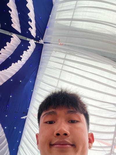 The image is a selfie taken from a low-angle perspective, looking upwards at the decorated ceiling of a tent or canopy. A young man, likely of East Asian descent, is featured prominently in the top right portion of the frame, appearing upside down from the viewer's orientation. He has short dark hair, an earring in his visible right ear, and looks directly into the camera with a neutral or slightly smiling expression. His eyes, nose, and part of his mouth are clearly visible.
The tent's ceiling is adorned with two primary fabric types. The upper section, where the man's head is largely positioned, consists of translucent white fabric panels with visible vertical folds, suggesting diffuse natural light or bright artificial lighting from above. The lower section is made of a deep royal blue fabric, embellished with decorative white ruffled fabric strips forming a wave-like or scalloped pattern. Numerous small, star-like lights, both white and blue, are embedded within the blue fabric, creating a festive atmosphere. Structural poles or framing are visible where the different fabric sections converge.
The scene depicts the interior of a decorated event tent, indicative of a celebration or gathering such as a wedding or party. The location context is Tay Son Commune, Vietnam. There is no visible text in the image.