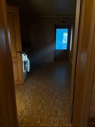 The image displays an interior space, likely a utility area or a small room, viewed from a slightly elevated perspective looking down. The walls are covered in square mosaic tiles, primarily in shades of light brown and beige, creating a textured, patterned surface. A prominent wooden frame borders the top and bottom of the immediate foreground, suggesting an opening or an architectural feature.

On the far wall, a rectangular window with a dark frame is visible. Through this window, a bright blue, indistinct mass can be seen, possibly indicating an external view of water or a vibrantly colored object. Distinct beams of sunlight stream through the window, sharply cutting through the dimness of the room and illuminating dust particles in the air. These light rays cast patterns onto the tiled walls and the surfaces below.

Beneath the window, a light-colored wooden cabinet or counter is present, featuring two cabinet doors. A dark, metallic object, possibly a key or a decorative pull, hangs from the right door. On the surface of this wooden unit, a white and grey appliance with a circular, translucent lid, resembling a small washing machine or a similar household device, is illuminated by the sunlight. A smaller, white rectangular object rests beside it.

The scene is static, without any human figures or discernible activity. The strong, direct sunlight indicates it is daytime, likely during clear weather. No visible text is present in the image. The environment, characterized by its tiled walls and wooden structures, suggests a functional space. The location is Shchelkovo, Russia.