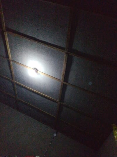 A low-angle shot shows a ceiling illuminated by a bare light bulb. The ceiling appears to be constructed with square panels, reinforced by wooden beams that create a grid pattern. The light bulb, centrally positioned, casts a strong glow, partially obscuring the ceiling's surface. Shadows and variations in light intensity suggest an uneven texture or slightly textured panels, while the overall scene is dark and somewhat grainy due to the low-light conditions.