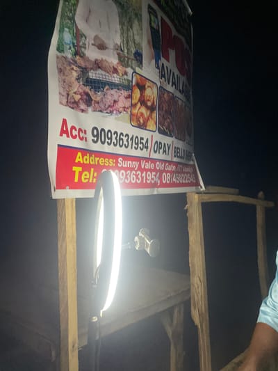 The image captures a nighttime scene, likely outdoors or in a dimly lit indoor setting, illuminated primarily by an artificial ring light positioned on the right. A large, vertically oriented banner or poster is prominently displayed on the left, advertising food items, specifically what appears to be skewered meat or kebabs, alongside other roasted or fried dishes.
A person's arm and shoulder, wearing a light blue garment, are partially visible at the top right corner. On the banner itself, a person in a white garment is depicted alongside the food. Above the ring light, a small camera or smartphone is mounted, suggesting an activity of recording or photography, possibly for advertising the food products.
The banner contains visible text providing business information:
- "AVAILAN" (likely part of "AVAILABLE")
- "Acc: 9093631954/OPAY / BELLO" (suggesting an account number for the OPay payment platform and a name)
- "Address: Sunny Vale Old Gate, FC Abuja" (indicating a location within Abuja, Nigeria)
- "Tel: 93631954, 08145022545" (providing contact phone numbers).
Wooden planks or scaffolding form part of a structure, possibly supporting the banner or acting as a stall framework. The overall scene, set in Galadimawa, Nigeria, depicts a food vendor operating at night, utilizing a ring light for illumination, possibly for a photoshoot or video, to promote their business.