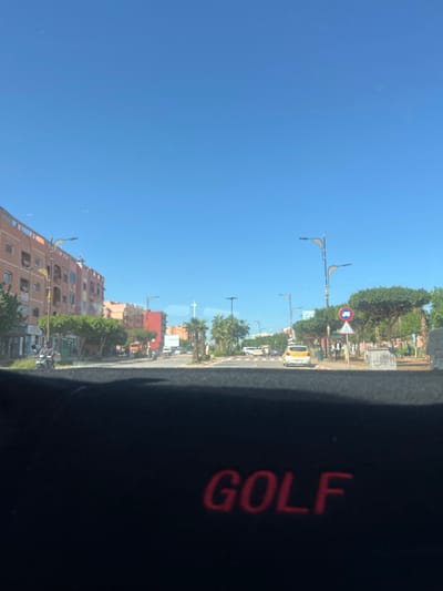 The image displays a wide urban street in Beni Mellal, Morocco, under a clear blue sky. The viewpoint is from inside a vehicle, evidenced by a black dashboard or mat with "GOLF" embroidered in red across the lower foreground.
On the left, a continuous line of multi-story buildings, predominantly peach-colored, features ground-floor commercial establishments. Green trees and streetlights line the sidewalk. Several motorcycles are visible, with one individual riding a scooter in the mid-ground. A red-colored building is situated further down the street.
To the right, a broad central median separates traffic lanes, populated with palm trees and other green trees with rounded canopies. Decorative streetlights are spaced within this median. A white and yellow taxi is observed in the rightmost lane of traffic. Multiple pedestrians are present within the median area. Traffic signs include a circular blue sign with white vehicle silhouettes, likely indicating traffic flow regulations, and a triangular warning sign depicting a pedestrian crossing. A large white vehicle, possibly a bus, is visible in the distance.
The paved roadway appears to accommodate multiple lanes of vehicular traffic. No specific interactions between individuals are discernible; general urban movement is depicted.