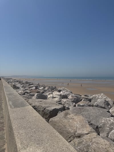 A bright, clear, sunny day is depicted at the beach in Wissant, France. The foreground is dominated by a concrete walkway and a large rock embankment. The walkway runs along the left side of the image, with visible brickwork beneath it. Beyond the walkway is a substantial pile of large, gray rocks, forming a breakwater.

The middle ground reveals a wide, sandy beach that stretches towards the sea. Several people are scattered across the beach, some walking, some standing, and some appearing to be in small groups. The water is calm and shallow, with ripples visible on the surface. The tide appears to be out, exposing a large expanse of wet sand.

In the distance, a hazy horizon line is visible, suggesting the presence of land or cliffs, and the ocean stretches out to meet the clear blue sky. The overall impression is one of a tranquil summer day at a coastal resort.