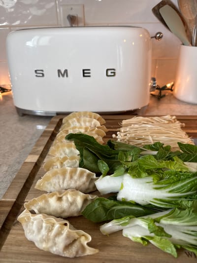 On a wooden cutting board, there is an arrangement of uncooked ingredients for a meal. The food includes a row of dumplings, a bunch of enoki mushrooms, and bok choy. A white Smeg-branded appliance is positioned to the left of the cutting board. The location is likely a kitchen or food preparation area, possibly in Orlando, United States.