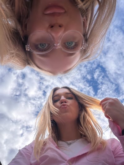 Two young Caucasian women are captured from a low-angle perspective, looking up at the camera against a backdrop of a cloudy sky. The woman in the foreground, on the lower right, has blonde hair and is wearing a pink button-down shirt over a white top. She is puckering her lips and holding a strand of her hair. The woman in the background, at the top of the frame, is also blonde and wearing glasses. Her head is upside down in the shot, with her eyes looking directly at the camera. The lighting suggests it is daytime. The location is specified as Dreiliņi, Latvia.