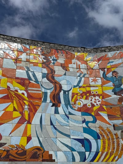 A medium shot looking up at a mosaic mural and a blue sky with white clouds, located in Gudauri, Georgia. The mural is made of individual square and rectangular tiles in various colors including red, orange, yellow, blue, grey, white, and brown. The mural features various stylized images including a person, a star, a machine, waves, wheat, and other abstract shapes. The mural is mounted on a grey stone wall.
