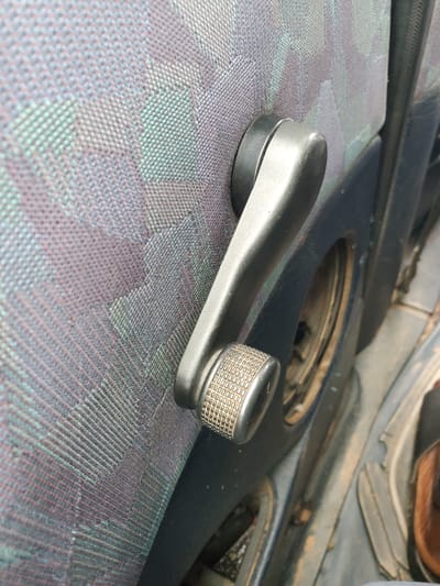 This is a close-up, eye-level, medium shot of a car's interior door handle and part of the door panel. The door panel is covered in a patterned fabric with shades of purple, green, and gray. A silver-colored window crank with a textured grip is mounted on the door panel, positioned horizontally. To the right of the crank, a dark-colored opening is visible, possibly where a speaker or other car component would be. The bottom right of the image shows a glimpse of a person's brown shoe and part of what appears to be a seat or floor mat. The lighting suggests an indoor or shaded environment. The scene is not located in Pankshin, Nigeria.
