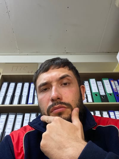 A man is taking a selfie in an office setting. He has dark hair, a beard, and is wearing a dark blue jacket with red accents. He is looking at the camera with his hand on his chin. Behind him are shelves filled with binders. There is also a box on the top shelf with red writing on it. The room appears to be lit by overhead fluorescent lights. The scene is consistent with an office environment in Luzhitsy, Russia.