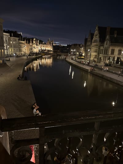 This is a nighttime, long-exposure photograph taken from a bridge overlooking a canal in Ghent, Belgium. The canal runs horizontally across the frame, reflecting the lights of the historic buildings lining its banks. On the left side of the canal, a cobblestone promenade is visible, with several groups of people sitting or walking along it. The buildings are characteristic of Ghent's architecture, with gabled roofs and illuminated windows. On the right side of the canal, more buildings are visible, also lit up, and a few boats are moored. The sky above is dark blue with streaks of clouds, and the water is dark, with shimmering reflections of the lights. The foreground of the image shows the dark, ornate railing of the bridge from which the photo was taken, with several padlocks attached to it, suggesting it is a "love lock" bridge. In the lower-left corner, a bright red object is partially visible. The overall atmosphere is tranquil and picturesque, capturing the beauty of Ghent at night.