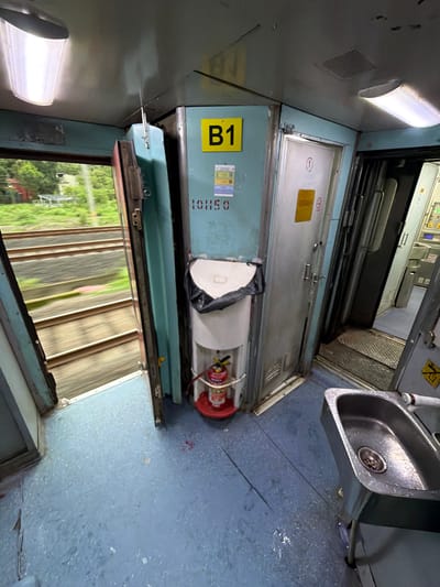 The image depicts the interior of a train carriage, likely a passenger compartment in Saphale, India. The view is from inside the carriage, looking towards the open doorway and a corridor. The floor is a light blue, speckled with some debris and what appears to be water or liquid stains. To the left, an open doorway reveals train tracks and blurred greenery outside, indicating the train is in motion.
Inside the carriage, a teal-colored wall features a yellow sign with the letter "B1". Below this, a number "101150" is stenciled in red. Adjacent to this is a white, wall-mounted water dispenser with a black bag covering the top of the container. A red fire extinguisher is visible at the base of the dispenser.
To the right of the dispenser is a silver door with a circular symbol and a yellow notice. Further into the carriage, another silver door is visible, partially open, leading to a different section. In the foreground on the right, a stainless steel sink with a faucet is positioned. The sink shows signs of recent use with water droplets. The overall environment suggests a functional, albeit somewhat worn, train interior. There are no people visible in the frame.
