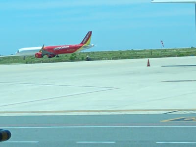 The image depicts an outdoor scene at an airport, possibly Cam Ranh International Airport in Bắc Cam Ranh, Vietnam, given the presence of an airplane with Vietnamese markings. In the foreground, a light gray tarmac is visible with white and red painted lines, indicating taxiways or runways. A portion of a dark-colored object, possibly a vehicle or part of airport infrastructure, is in the lower left corner.
In the midground, two airplanes are present on the tarmac. The primary subject is a red airplane with yellow accents, bearing the "Vietjet Air" logo on its tail. This aircraft appears to be moving or stationary on the tarmac. Behind it, a white airplane is partially visible. The background consists of a green, grassy area with scattered vegetation. Further in the distance, a windsock with red and white stripes is visible on a pole, suggesting windy conditions. A bright orange traffic cone is positioned on the tarmac in the right midground. The sky is clear and light blue. The overall impression is a typical airport operational environment on a clear day.