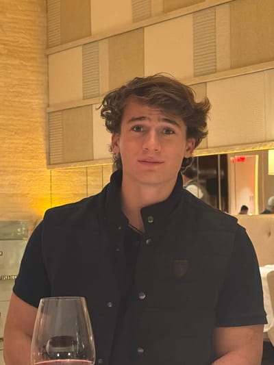 A young man with curly brown hair looks at the viewer. He wears a dark blue button-up shirt. The background includes a textured wall and part of a wine glass. The image appears to be taken indoors, possibly in a restaurant, judging by the ambient lighting and decor.