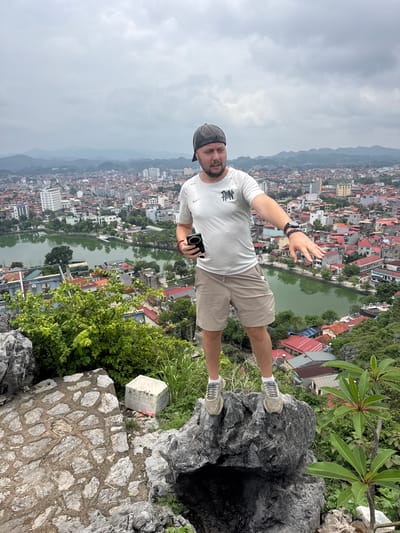 A man stands on a rocky outcrop, pointing towards a city below. He is wearing a white t-shirt, beige shorts, a baseball cap, and athletic shoes. In his right hand, he holds a black smartphone. The city below is dense with buildings, many featuring red tile roofs, and a winding river or lake runs through it. The sky is overcast, and a range of hills or mountains is visible in the background. The man appears to be a tourist in Tam Thanh Ward, Vietnam, possibly at a viewpoint overlooking the town.