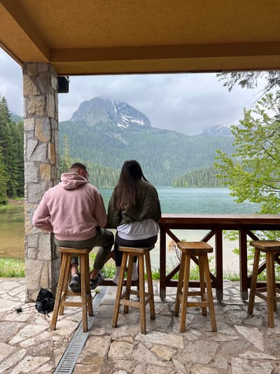 This image captures a serene outdoor scene from a sheltered viewpoint, likely a cafe or observation deck, overlooking a magnificent natural landscape. Two individuals are seated on high wooden bar stools, their backs to the camera, engrossed in the view.
The person on the left is wearing a pink hoodie and olive green pants, with short dark hair. The person on the right has long dark hair and is dressed in a dark green jacket over a white top and dark leggings. They are looking out towards a large, light green-blue lake, which is bordered by dense green forests. Beyond the forests, rugged mountains with visible patches of snow rise under an overcast sky.
The immediate foreground features a textured stone floor made of irregular flagstones. A sturdy wooden railing system separates the seating area from the expansive view. To the left, a stone wall or pillar is visible, capped by a yellow-stuccoed overhang or ceiling. Sparse green tree branches hang into the frame from above. On the floor near the stools, a black bag and a metal drainage grate can be seen.
The atmosphere is peaceful and contemplative, with the cloudy weather adding a soft, diffused light to the dramatic mountain and lake scenery. The time of day is daytime, though the overcast conditions prevent bright sunshine. No visible text is present in the image. This picturesque setting is located in Pitomine, Montenegro.