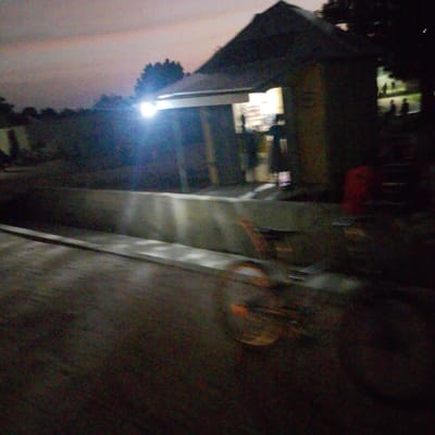 This is a blurry, low-light photograph taken at dusk in Gulak, Nigeria. The scene depicts an outdoor setting with a simple structure that appears to be a shop or food stall. The building has a dark, sloped roof and an open front, with interior lights illuminating shelves and what might be products. Silhouettes of a few people are visible inside and outside the structure, suggesting it is open for business.
In the foreground, a blurred object, possibly a bicycle, is in motion, indicated by the streaky motion blur of its wheels and frame. The ground is paved and appears to be a road or pathway. A concrete barrier runs horizontally across the mid-ground, casting faint reflections from overhead lights. The background shows a dark, overgrown area with trees and vegetation. The sky is a hazy gradient of purplish-grey, consistent with twilight. The overall impression is of a modest, functional establishment in a rural or semi-rural Nigerian community at the end of the day. No visible text is discernible due to the image quality and lighting conditions.