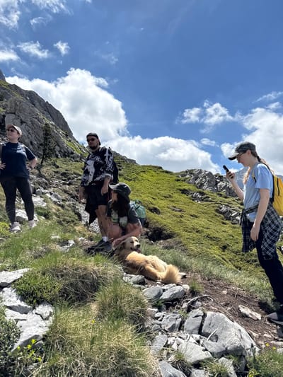 In Jošanica, Montenegro, a group of four individuals and a dog are seen on a grassy, rocky mountainside under a bright blue sky with scattered clouds. The person on the far left, wearing a dark t-shirt and leggings, stands with hands on hips. To their right, a person in a black and white long-sleeved shirt and shorts stands, with another person sitting beside them. This seated individual, wearing a light-colored top, is partially obscured by a golden retriever lying down in front of them. The person on the far right, wearing a light blue t-shirt and a plaid shirt tied around their waist, is looking at their phone and holds a yellow backpack. The environment is characterized by steep, rocky terrain and sparse vegetation.