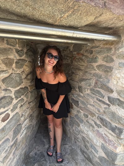 A woman stands in a stone alcove, likely part of the Walls of Kavala, Greece. She is wearing a black off-the-shoulder dress, sunglasses, and sandals. She has curly brown hair and tattoos on her arms and legs. Her hands are clasped in front of her, holding a dark-colored object, possibly a phone. Two metal pipes are visible above her head, supported by the stone structure. The walls are constructed of rough, grey stones of varying sizes, cemented together. The lighting suggests it is daytime, with sunlight casting shadows within the alcove.