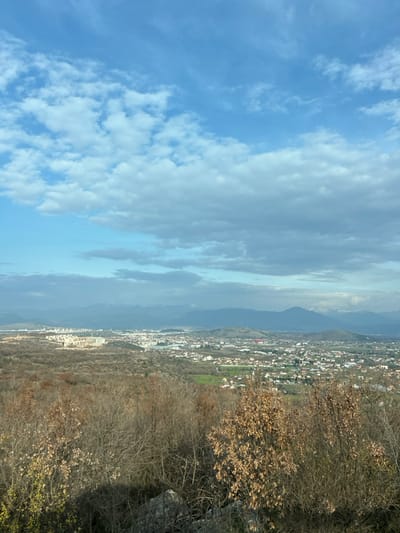 This image presents a panoramic landscape view captured from an elevated position within Gornji Kokoti, Montenegro. The foreground is dominated by a dense cluster of deciduous trees and shrubs with bare, dark branches and scattered remnants of dry, light brown foliage, indicative of late autumn or winter. Rocky terrain is visible at the immediate base of this vegetation.

The midground features an extensive urban settlement, likely encompassing parts of Gornji Kokoti and surrounding areas. This settlement consists of numerous residential and commercial buildings of varying sizes and colors, with a concentration of larger, lighter-toned multi-story structures visible on the left side of the urban sprawl. Green fields and cultivated land are interspersed throughout and bordering the built environment.

The background reveals multiple layers of mountains extending across the horizon. Distant peaks display visible snowcaps, suggesting higher altitudes. The sky, occupying the upper half of the frame, is partly cloudy, with bright blue patches interspersed with white, scattered cumulus clouds and broader, grayish stratocumulus formations. The lighting suggests daylight conditions, with shadows indicating a sun position to the left of the observer. No individuals or active human interactions are depicted within the scene.