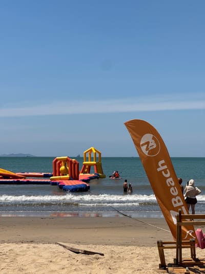 An aerial view of an inflatable water park situated in the ocean off the coast of Quy Nhon Ward, Vietnam. The park is composed of various interconnected inflatable structures, including slides and climbing walls, in vibrant shades of orange, yellow, red, and blue. Several individuals can be seen enjoying the water park, with some on the inflatables and others in the water. The scene is set against a clear blue sky and the vast expanse of the ocean, with a sandy beach in the foreground. An orange flag with a logo and the word "Beach" written vertically is prominently displayed on the right side of the image, partially obscuring the view of the water park. The flag appears to be a marker or advertisement for the beach area. The water is a deep blue, and gentle waves are lapping at the shore. Distant landmasses and ships are visible on the horizon.