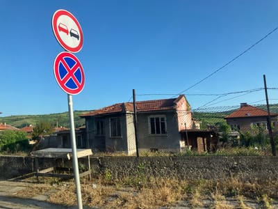 The image depicts a street scene in what appears to be a rural or semi-rural area in the city of Biaga, Bulgaria. The foreground contains two circular road signs mounted on a single pole: the upper sign indicates "no overtaking" with two cars in red and black, while the lower sign indicates "no parking" with a red circle and blue X. To the right of the sign, there is a weathered concrete fence with overgrown vegetation. Beyond the fence, there are several dilapidated buildings with terracotta roofs and wire fences. The landscape in the background features a rolling green hill under a clear blue sky. The scene appears deserted, with no people visible.
