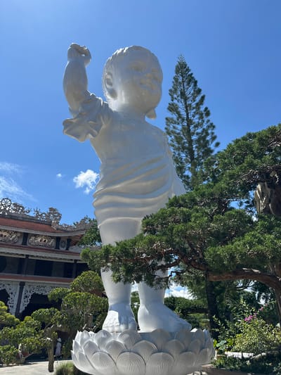 The image, captured in Thôn 5, Vietnam, features a large, white statue of a baby in a playful pose against a bright blue sky. The statue appears to be made of smooth, polished material and is detailed with flowing drapery. The baby is depicted with a round face, chubby arms, and delicate fingers, with one hand extended forward as if reaching for something. The statue rests on a base with a decorative, petal-like pattern.
Surrounding the statue are lush green trees, some with intricate, sculpted branches that frame the baby figure. In the background, traditional Vietnamese architecture is visible, characterized by ornate rooflines, decorative carvings, and intricate latticework. These architectural elements suggest a temple or a significant cultural site.
The lighting indicates it is daytime, with clear, sunny weather as evidenced by the bright sky and the shadows cast. The overall scene evokes a sense of peace and possibly spiritual contemplation, given the religious architectural context and the serene depiction of the baby. The image is oriented vertically, emphasizing the height of the statue and its surrounding elements. No visible text is present in the image.